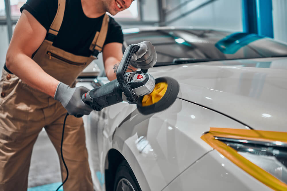 Professional auto detailer hand holding rotary polisher while polishing paint surface of shiny white car.