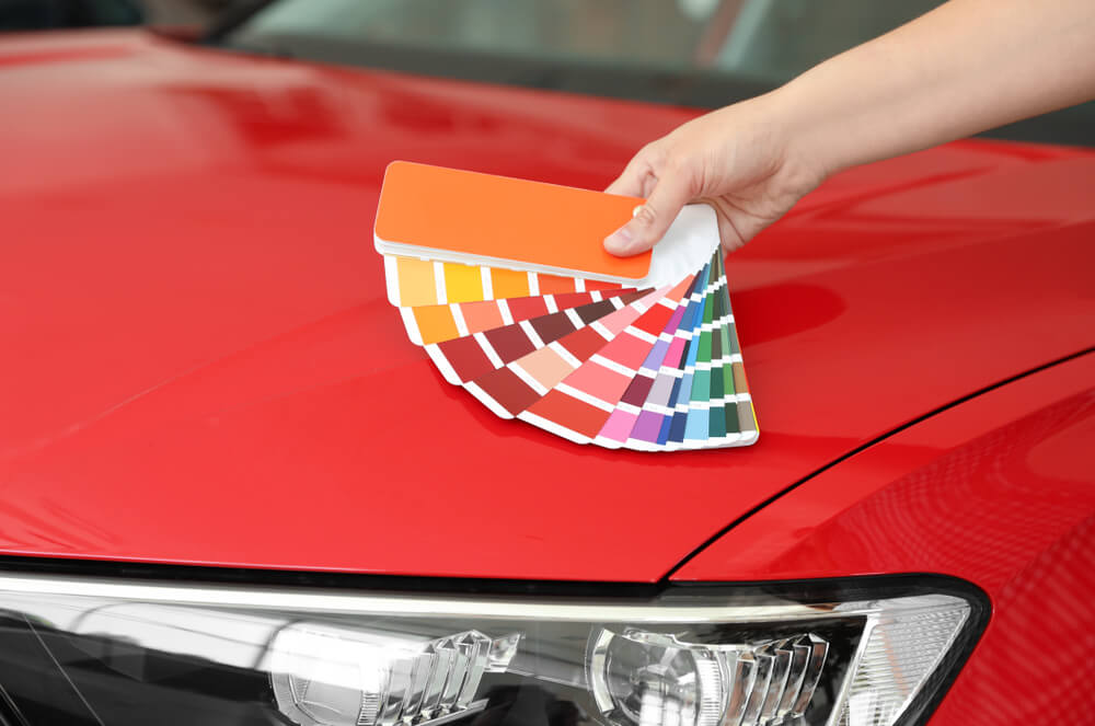 Young woman holding palette with color samples near red car.