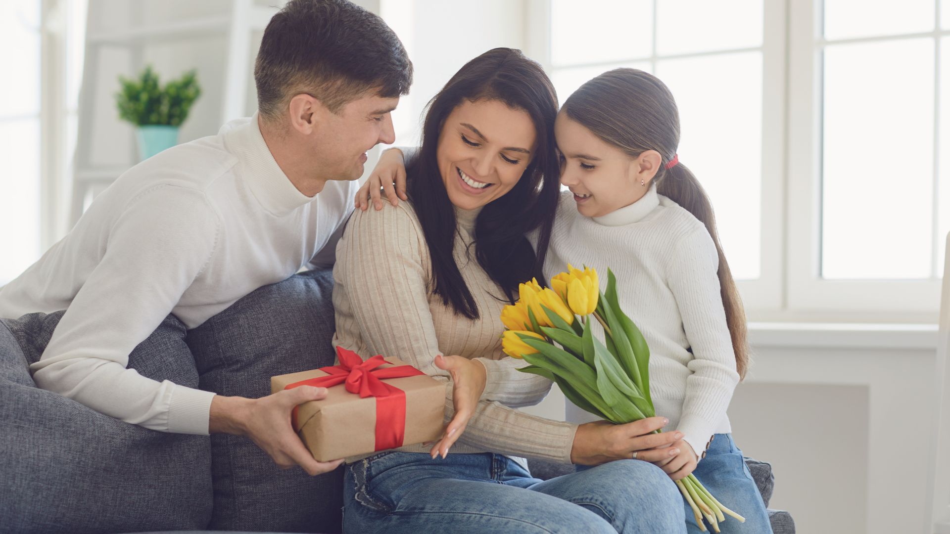 a man and child give a woman a gift for Mother's Day while sitting on a couch.